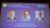 A screen displays the portraits of the laureates of the 2019 Nobel Prize in Chemistry (L-R) John B. Goodenough, M. Stanley Whittingham, and Akira Yoshino