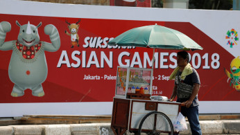 A food vendor pushes his cart past a sign promoting the upcoming Asian Games in Jakarta
