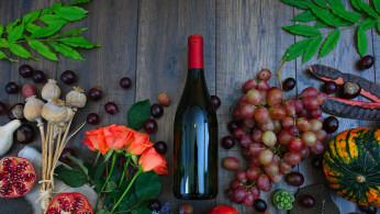 Wine bottle beside grapes roses and several fruits on brown wooden surface