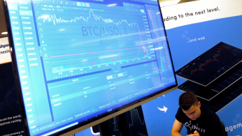 A man works beneath a display showing the market price of Bitcoin on the floor of the Consensus 2018 blockchain technology conference
